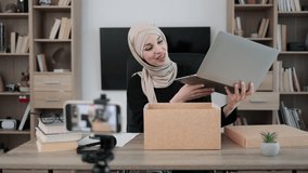 Pleasant arab woman filming video on modern phone camera while opening parcel box with new wireless laptop. Concept of people, technology and blogging.