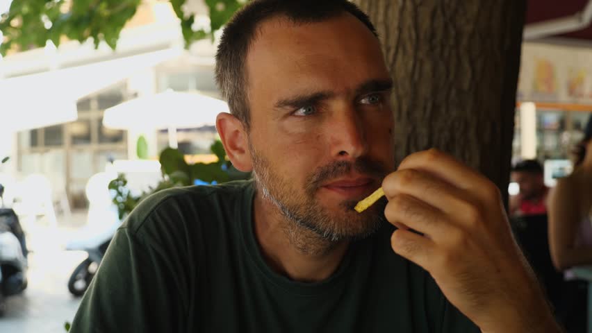 A handsome middle-aged man eats French fries, fried potatoes sitting in an outdoor cafe. The guy is enjoying his fries, looking at the camera. The concept of unhealthy and fast food | Shutterstock HD Video #1099445203