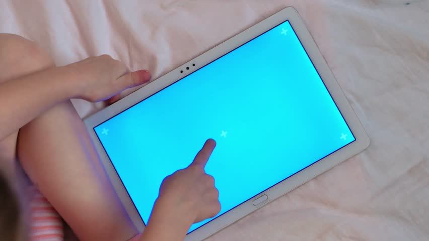 A little girl runs her finger on a tablet with a blue chromakey screen playing video games in a white bed, top view.