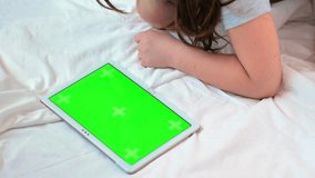 A little girl in a white bed is looking at a tablet teaching lesson or cartoon lying on her stomach screen with a green chrome key screen. Wearily lays his head on the bed.