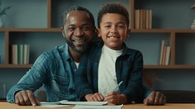 African American father dad with little son kid child boy schoolboy schoolchild schoolkid sit at table after homework writing home learning education looking to camera waving hello greeting smiling
