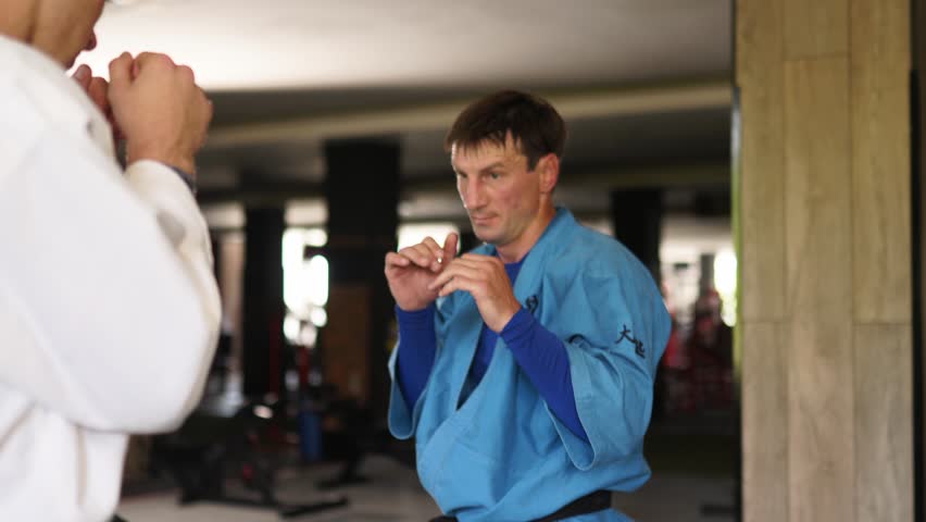 Two martial arts men in kimonos practice kicks and punches. Male fighters wearing kimono training in gym. Kick boxers jumping and practicing kicking and punching actions indoors. People and sport  | Shutterstock HD Video #1099460871