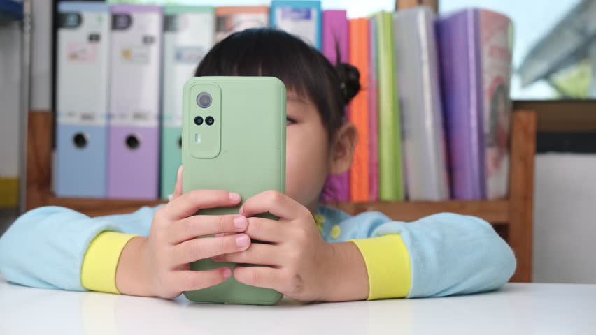 Cute little girl holding smartphone looking at screen smiling using application, playing online games, watching cartoons in living room at home. Children and modern technology concept | Shutterstock HD Video #1099462193