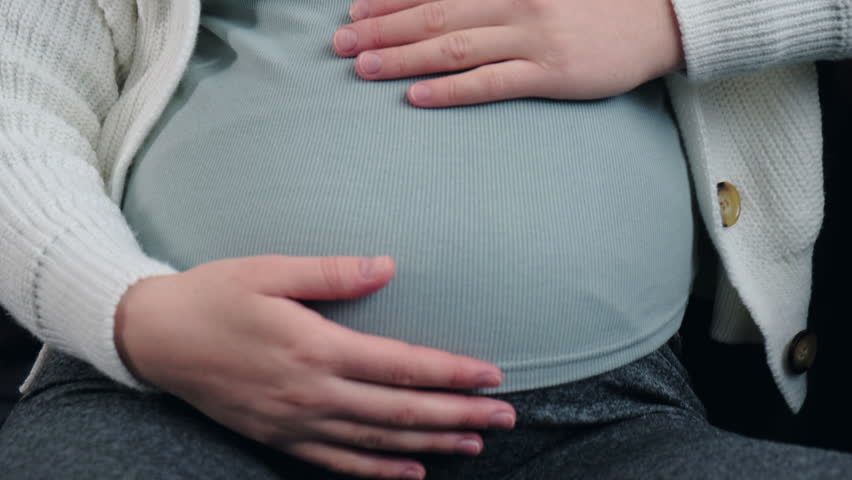 Selective focus of upset young pregnant woman sitting on sofa in living room at home, feeling discomfort ache in neck and massaging shoulders, tensed muscles during pregnancy. Fibromyalgia concept