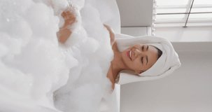 Vertical video of smiling biracial woman blowing foam in bathtub in bathroom. Health and beauty, leisure time, domestic life and lifestyle concept.
