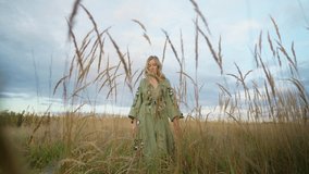 View of beautiful blond woman, walk through the wheat field at dawn, high plants stems, autumn comes. Romantic, enigmatic silhouette, green embroidered dress. Sunset out of the city. High quality 4k