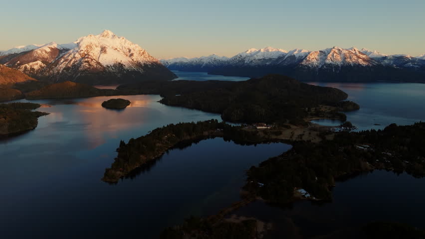 A wide drone flight panning from left to right, over the Nahuel Huapi lake with a view of the sunlit Tronador mountain at Bariloche, Argentina during sunrise