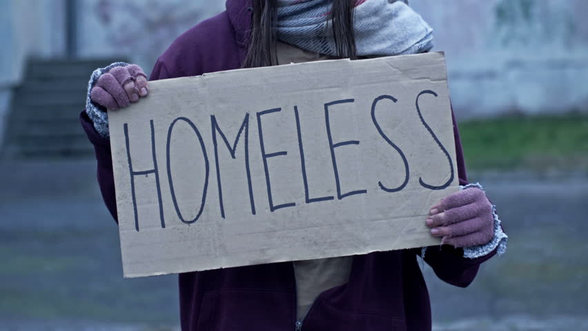 An unkempt-looking young woman sits with a begging cup and holds a handwritten HOMELESS poster.