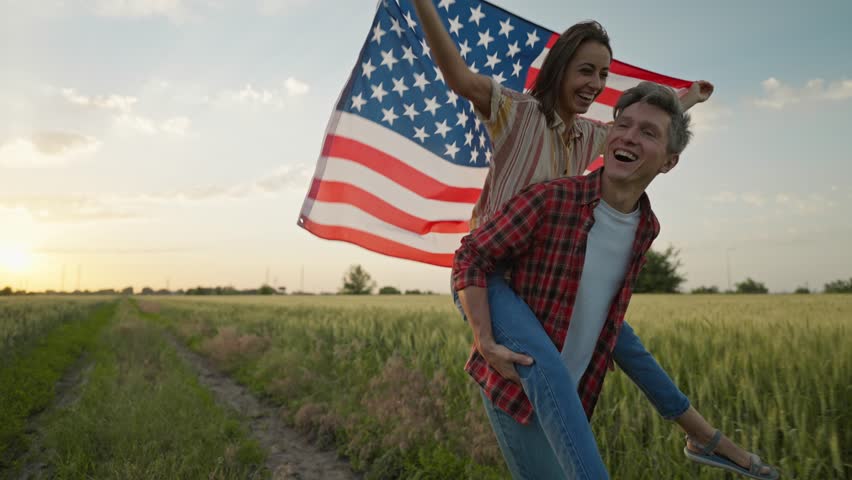 4k slow motion of happy patriotic couple celebrating fourth of july with national flag running in rural lardmark at sunset. 4th of July. USA independence day celebrating with American flag.