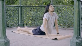 Young hippie latin man with long hair doing the cobra pose on a yoga mat in a wooded area surrounded by a railing. 4k video