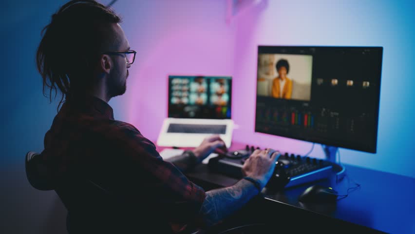 Young man in glasses working on video editing on computer doing montage and color correction. Professional man hand making edits on a special panel. Concept of creating video content. | Shutterstock HD Video #1099480401