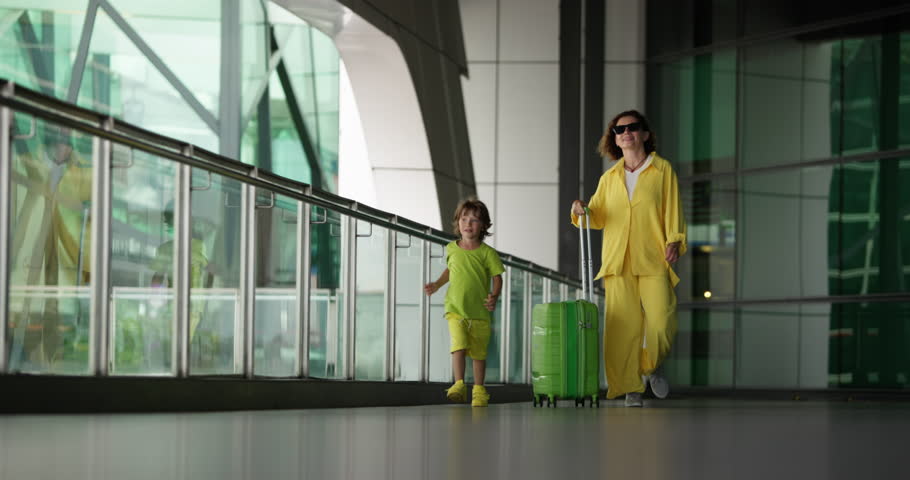 Young, modern mother with son walks through airport after landing and dances, rejoicing at arrival on vacation. Mother and son are dancing, jumping in airport building for joy. Mom gives son high five Royalty-Free Stock Footage #1099482721