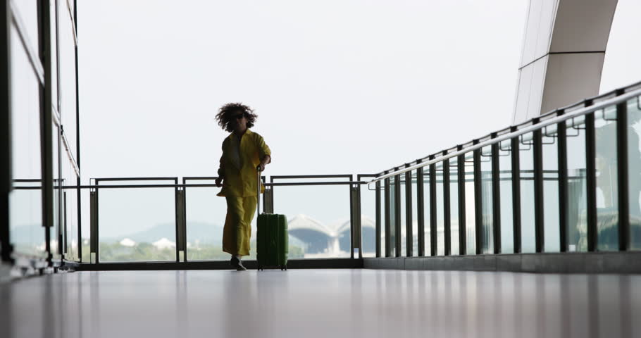 Low angle shot of curly-haired woman in a yellow outfit hopping, dancing and shaking her head from side to side, rejoicing at her arrival at the airport. A woman walks, dances and enjoys her vacation. Royalty-Free Stock Footage #1099482743