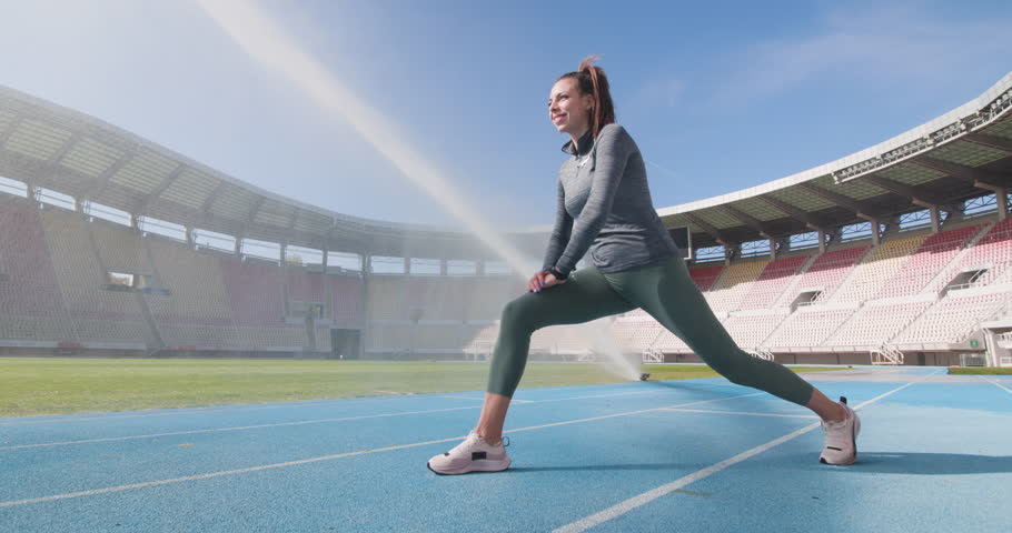 Sports runner girl stretching and warming up on synthetic track at open field generic stadium. Water irrigation in the backgorund.