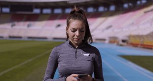 Female runner girl walking forward. Checking her phone. Open field generic stadium in the background. Handheld video.