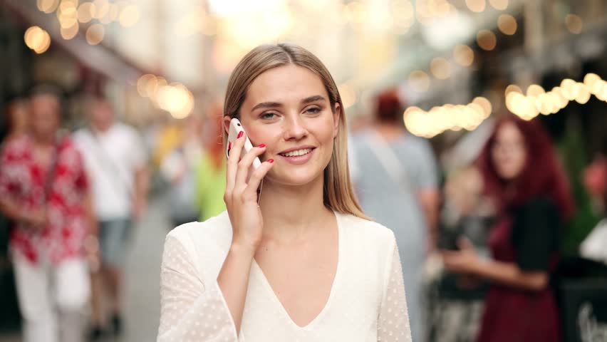 Slow motion front view of a young beautiful blonde woman in a white dress walking and calling on phone in the street. | Shutterstock HD Video #1099487263