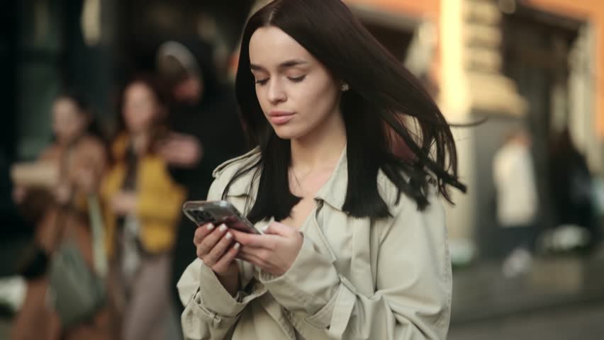 Smiling beautiful woman wearing trendy glasses walks down the old city street and uses her phone. Pretty girl in white shirt walks down the street looking at her mobile phone | Shutterstock HD Video #1099487265
