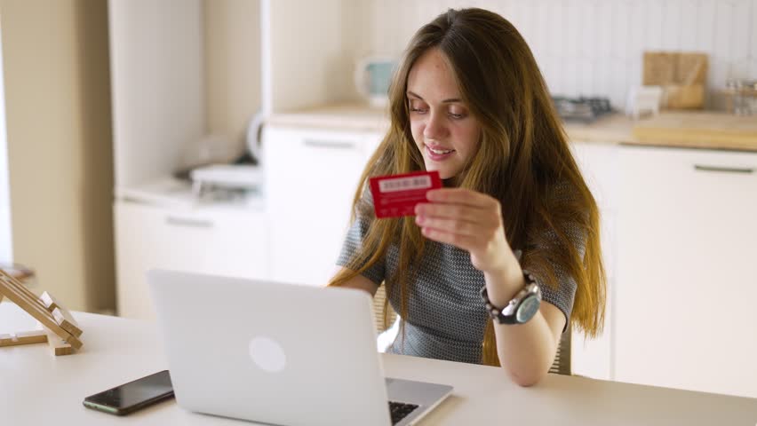 Cheerful girl makes purchases in the online store at home. Internet sale. Shopping with a card and a laptop. Shopping online. E-commerce sitting at home at the desk. Credit card in the hands of a girl | Shutterstock HD Video #1099487951