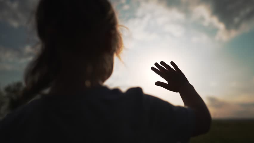 Girl waves her hand at sun. Religion is a joy to God. Silhouette of a child at sunset in the park. Girl raises her hand to the sky at sunset. Children's palm in the rays of the sun. Religion concept Royalty-Free Stock Footage #1099487975