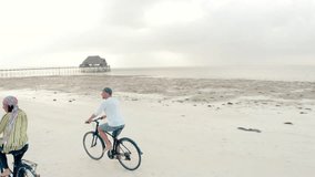 4K video couple of people on bicycles going along sandy beach at sunset with pier in background. Environment, healthy lifestyle, spending time outdoors concept. Zanzibar, Tanzania.