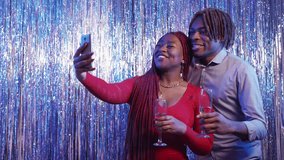 Mobile greeting. Beloved couple. Party celebration. Joyful black man and woman taking video with champagne on smartphone posing shimmering cascade curtain background.
