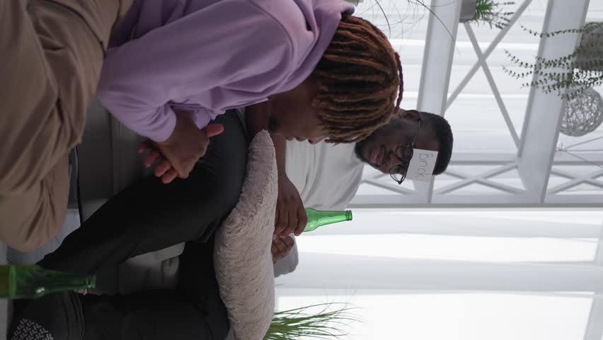 Vertical video. Home entertainment. Relaxed male friends. Happy black men playing game guess who with pizza and beer sitting light room interior.