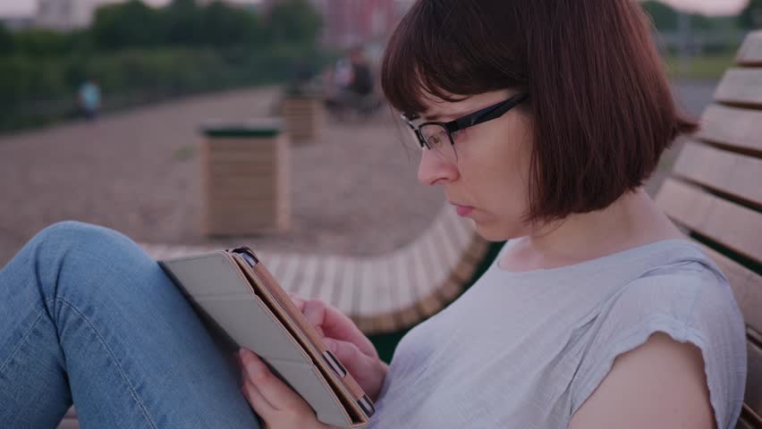 Side view of a female freelancer with bob haircut glasses working on a digital tablet while sitting on a chaise lounge in an early morning public park, copy space | Shutterstock HD Video #1099493727