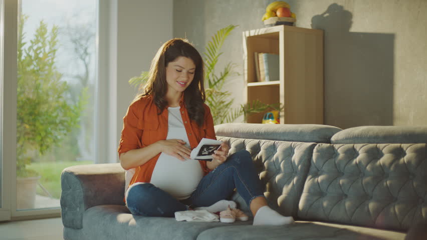 Young pregnant woman smiling and touching her belly by sitting on the sofa while looking at her ultrasound report Royalty-Free Stock Footage #1099493917