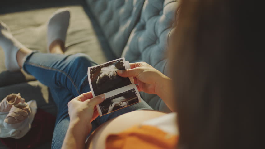 Close-up back view of a pregnant woman watching her baby ultrasound report and touyching her belly while sitting on sofa at home Royalty-Free Stock Footage #1099494017