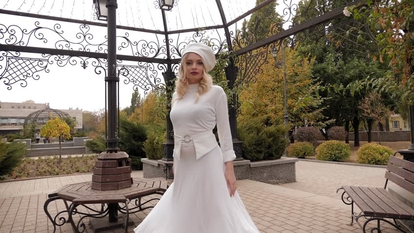 A gorgeous young woman in an elegant white dress and a white hat poses in a city Park in a metal wrought-iron gazebo.