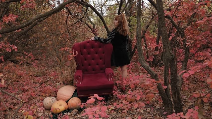 A luxury model poses in the woods in autumn next to a red antique chair and pumpkins.
