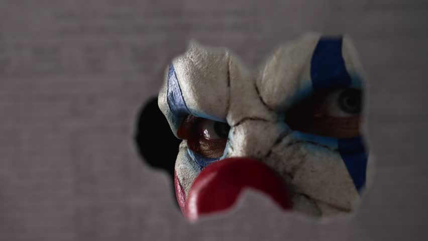 A scary clown face peeking through a hole in the partition. Close-up. The face of a man wearing a creepy clown mask peering at his victim through the hole. The creepy clown maniac | Shutterstock HD Video #1099502091