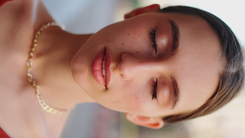 VERTICAL VIDEO: Сlose-up view of a beautiful girl with brown hair opening her eyes and smiling to the camera on city background.Portrait ofromantic girl smiles broadly on urban background.