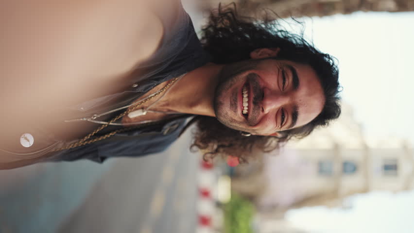 VERTICAL VIDEO: Young italian guy with long curly hair and stubble takes selfie on mobile phone while standing on the street. Stylish man posing smiling at the camera of his smartphone Royalty-Free Stock Footage #1099502883