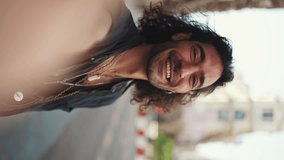 VERTICAL VIDEO: Young italian guy with long curly hair and stubble takes selfie on mobile phone while standing on the street. Stylish man posing smiling at the camera of his smartphone