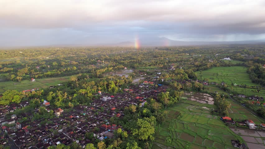 Small dense build Balinese village and rice fields seen down, light rainy clouds in the air with a short rainbow below them. Aerial shot in morning hour, typical countryside at north of Ubud town | Shutterstock HD Video #1099504321