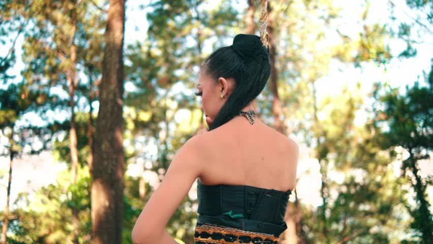 A slow-orbiting shot from behind a woman in a green skirt and black corset, wearing a traditional golden headpiece. She poses gracefully in a sunlit pine forest.