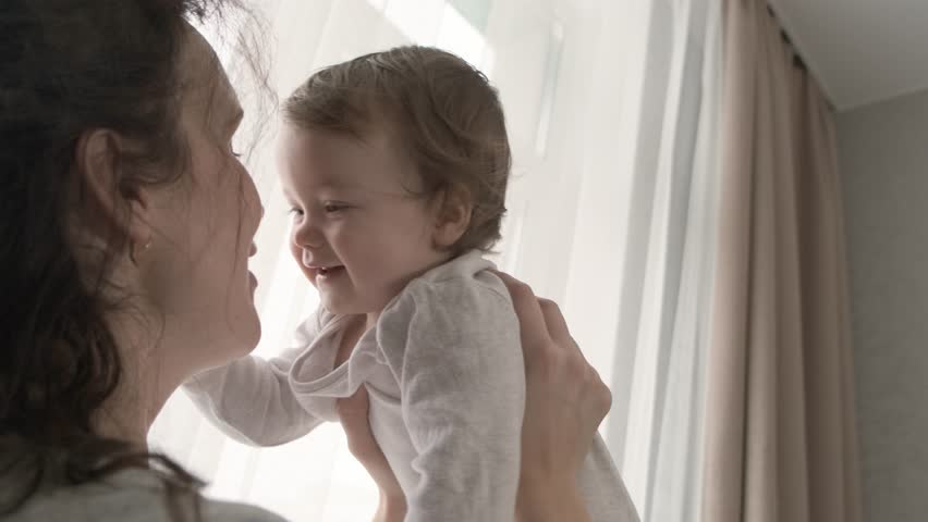 Mother and her baby girl having fun and playing at home. Little kid 1 year old play with his mom arms at home near a big window with sunlight | Shutterstock HD Video #1099512475