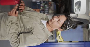 Vertical video of biracial female car mechanic using tablet and smiling. Working in car repair shop and running small feminine business concept.