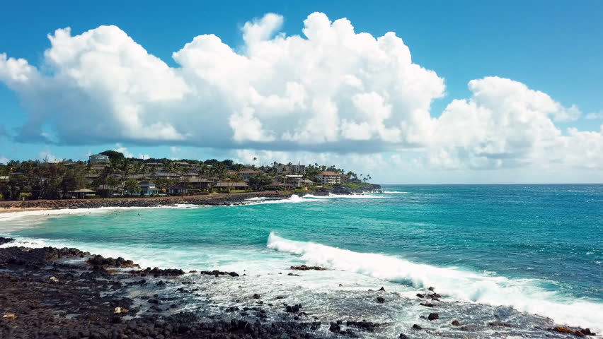 Aerial shot along Poipu Beach; featuring rocky surf, sand beaches and beachside residences.