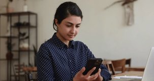 Positive millennial Indian business freelance woman reading, typing text message on mobile phone, using app on Internet, chatting, smiling at good news, looking away, thinking at office work table