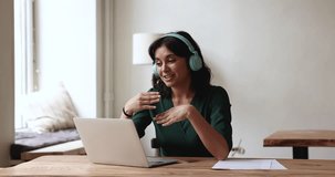 Engaged Asian businesswoman in big wireless headphones talking to client on video call at laptop, speaking, gesturing, smiling, sitting at workplace table with desktop, working on communication