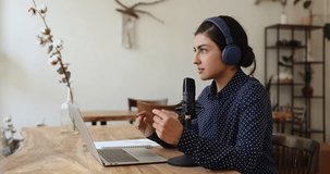 Busy focused Indian influencer, blogger in wireless headphones giving live broadcasting, speaking at audio equipment microphone, laptop in sound studio, recording podcast, talking to audience