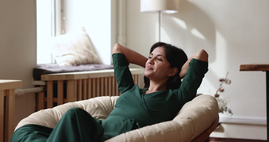 Happy joyful relaxed Asian woman resting in comfortable round armchair, holding head, touching nap, taking deep breath, enjoying leisure, comfort at home, looking away, thinking, dreaming, smiling Royalty-Free Stock Footage #1099514571