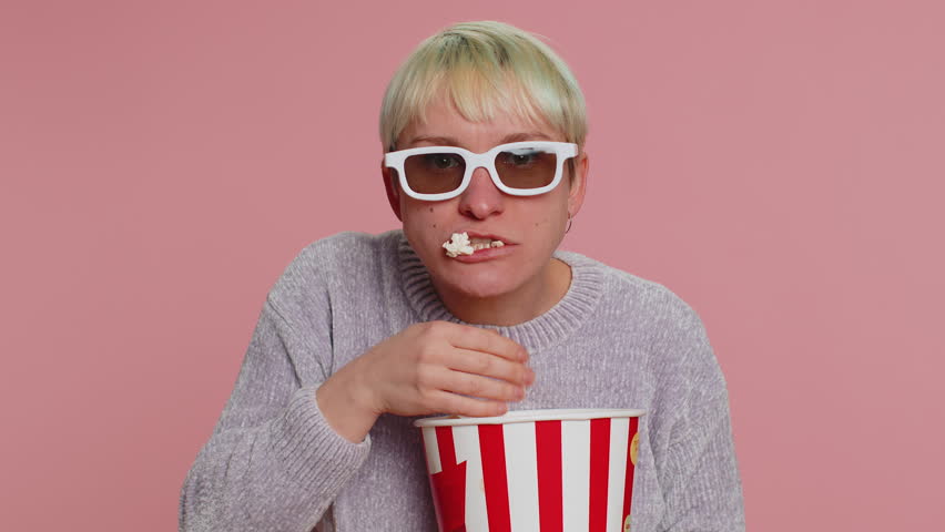 Excited woman in 3D glasses eating popcorn, watching interesting tv serial, sport game, film, online social media movie content. Young girl enjoying domestic entertainment on pink studio background | Shutterstock HD Video #1099518375