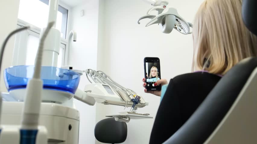 A young blonde in a dental chair communicates via the Internet from a smartphone. | Shutterstock HD Video #1099518961