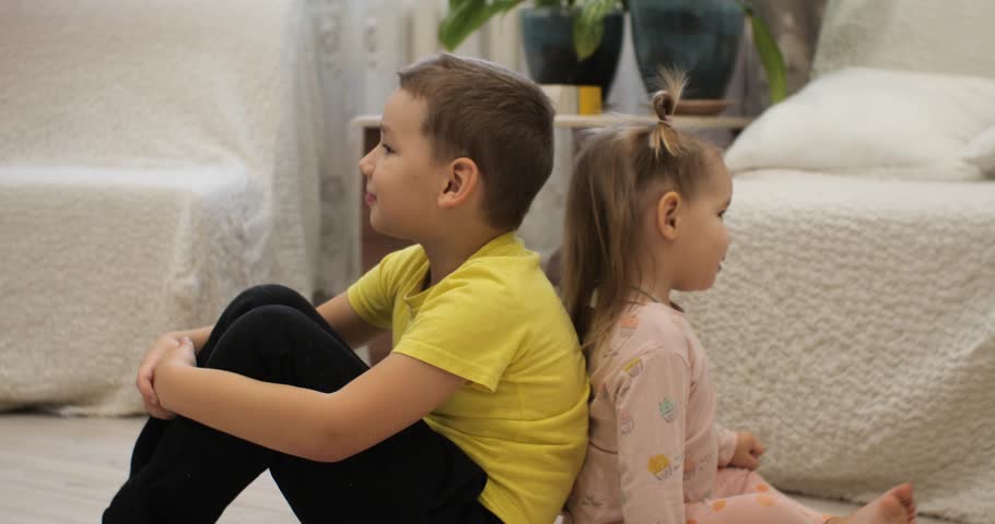 A boy and a girl sit back to back on the floor and smile thoughtfully. The children leaned on each other sitting on the floor of the living room. Family concept with children. | Shutterstock HD Video #1099520141