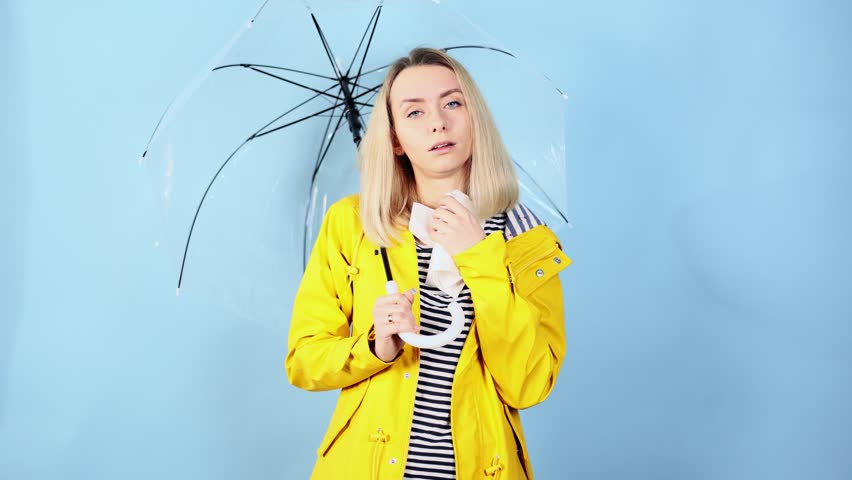 Sick young blond woman in yellow raincoat blowing running nose sneezing and coughing on isolated blue background Unhealthy female under umbrella getting flu virus symptom Cold and fever concept | Shutterstock HD Video #1099522677