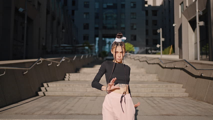 Young woman with tattoo on belly dances hip-hop on stairs against building. Dancer wearing black blouse and pink trousers shows energetic moves slow motion | Shutterstock HD Video #1099526421