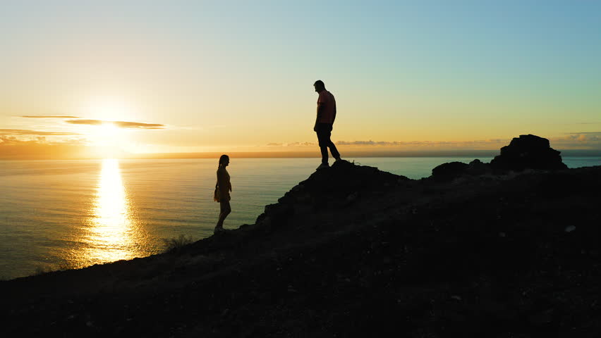 Couple of travelers climb the mountain during sunset and look at ocean sunset. Man gives a helping hand to womn. Travel vacation adventure teamwork concept.